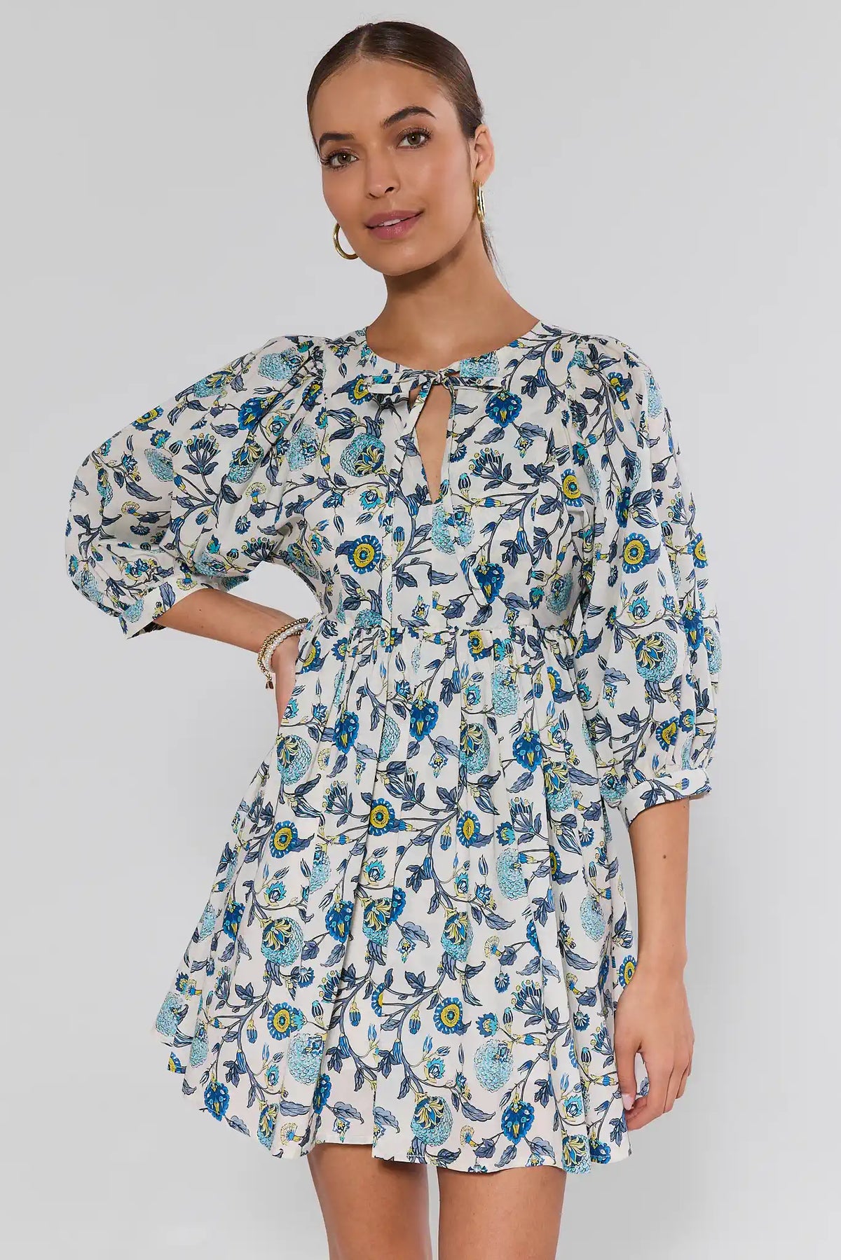 Model wearing Paige Blue Floral Print Short Dress with puffed sleeves, tiered design, and fitted waist, highlighted by gold hoop earrings and bracelets.