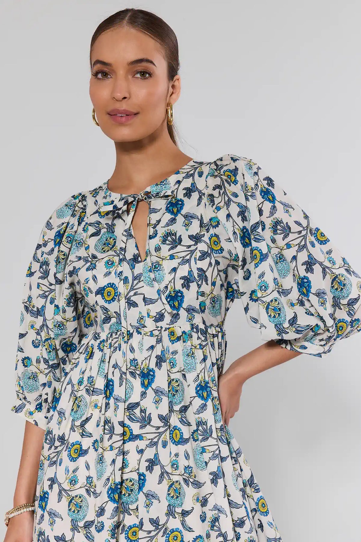 Woman wearing Paige Blue Floral Print Short Dress with puff sleeves, A-line silhouette, keyhole neckline, and gold hoop earrings, hand on hip, hair pulled back.