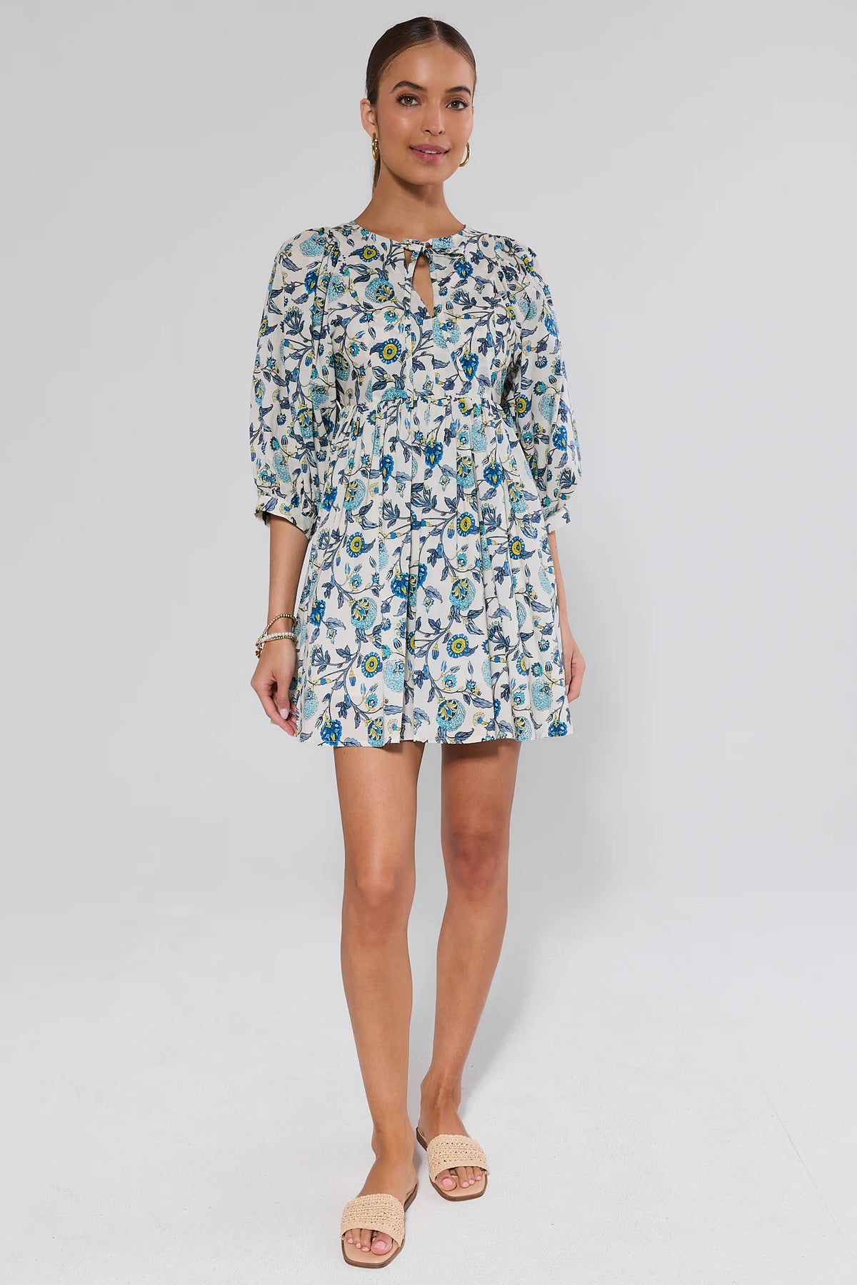 Paige Blue Floral Print Short Dress worn by a dark-haired woman, featuring a keyhole neckline, tiered design, and puff sleeves, paired with beige woven sandals.