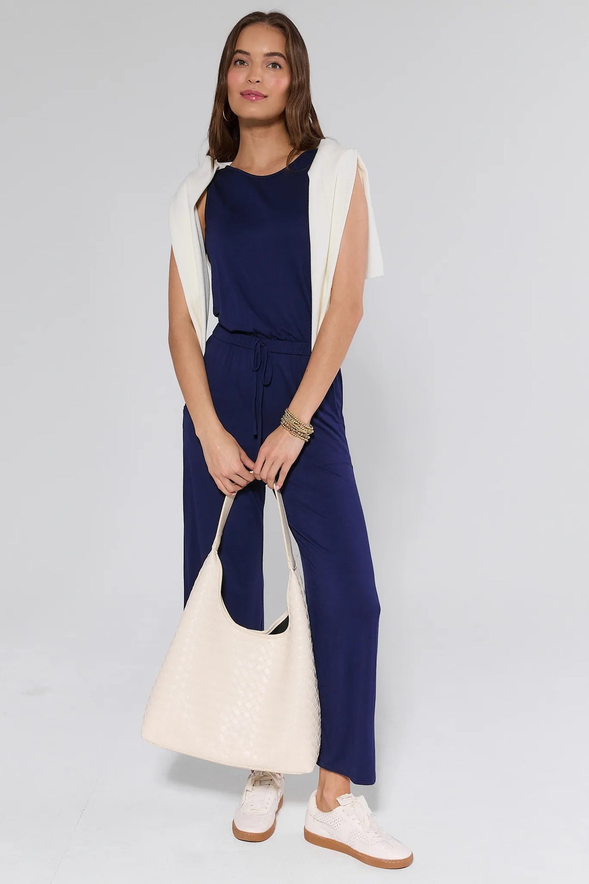 Woman wearing a navy jumpsuit with a white top and holding a white bag on a light gray background
