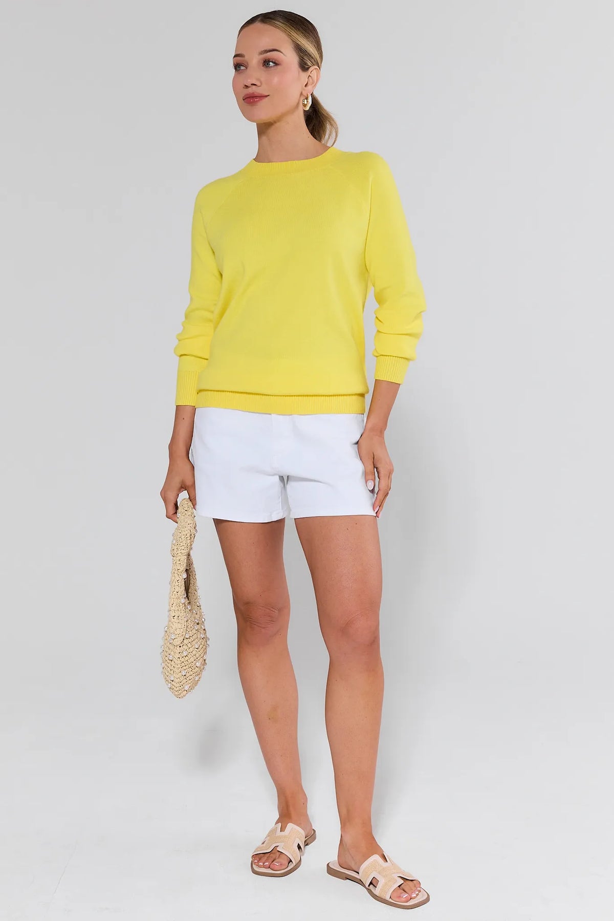 Woman models Jane White Finished Hem Shorts, styled with a yellow knit sweater, beige sandals, and a woven tote, capturing a relaxed, casual spring/summer look.