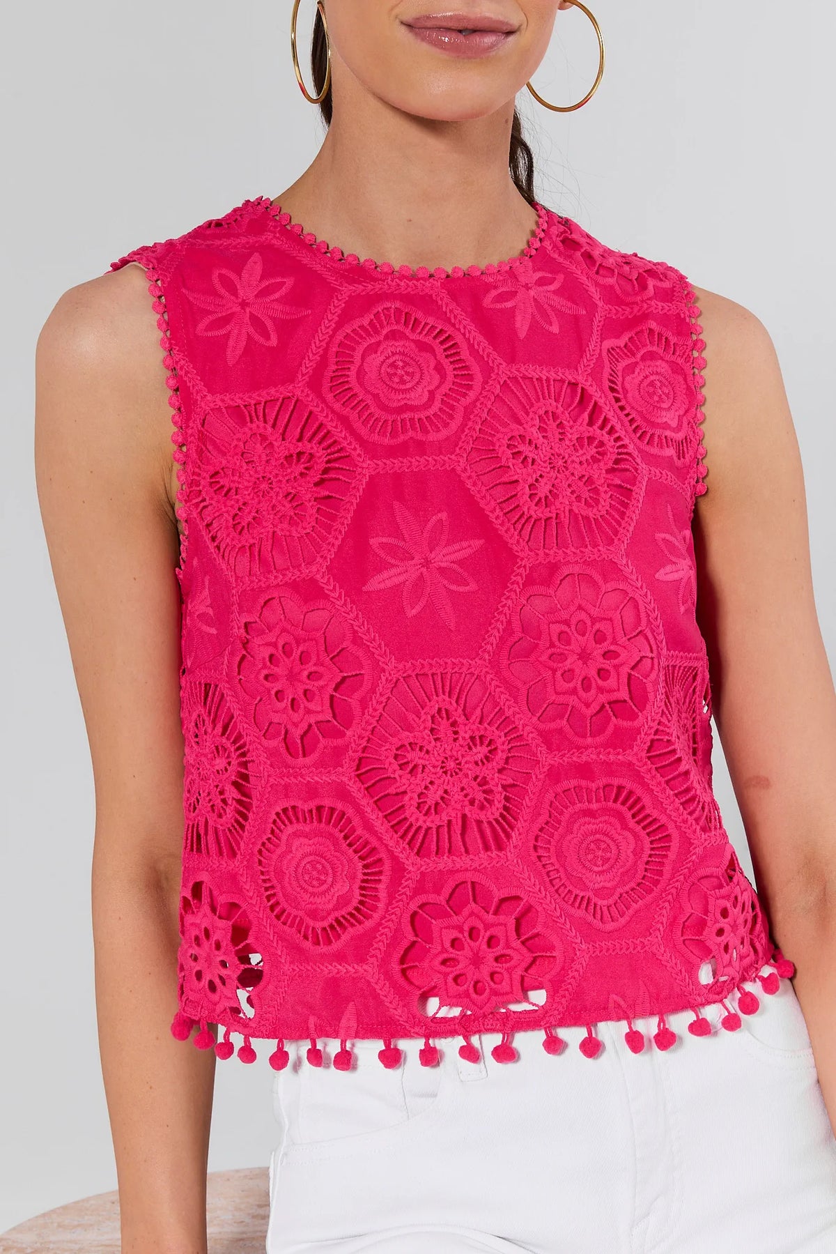 The Arden Pink Eyelet Top features a sleeveless, hot-pink crochet design with interlocking hexagonal panels and pom-pom trim, styled with white bottoms.
