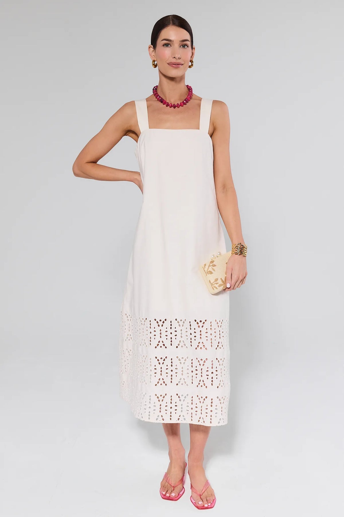 Model wearing the Melody Bone Eyelet Midi Dress with a square neckline, accessorized with a fuchsia necklace, gold jewelry, beige clutch, and pink sandals.