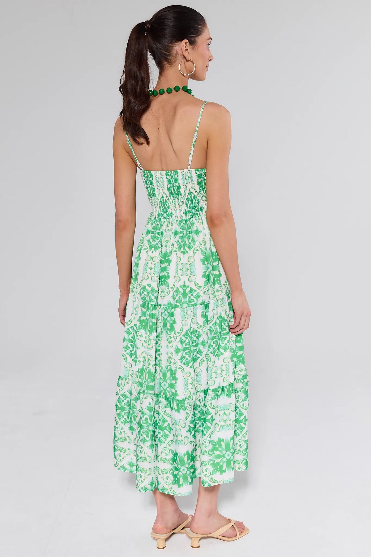 Woman in Lori Green Printed Midi Dress with spaghetti straps, smocked bodice, tiered skirt, styled with beige sandals, gold hoop earrings, and a green bead necklace.