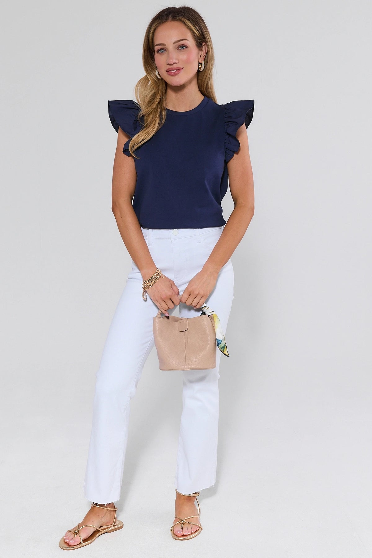 Woman wearing a navy top with ruffled sleeves and white pants, holding a beige handbag on a light gray background.