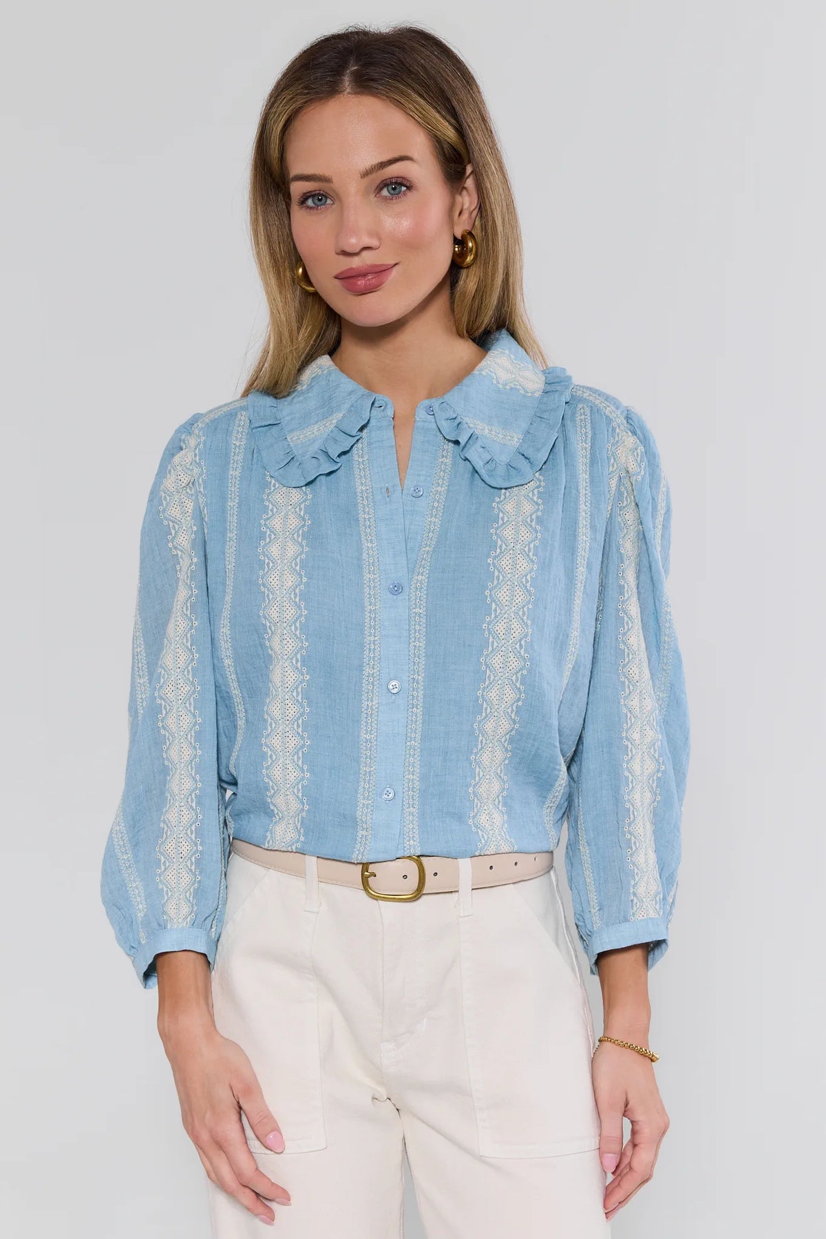 Woman wearing a light blue blouse with ruffled collar and white pants on a plain background