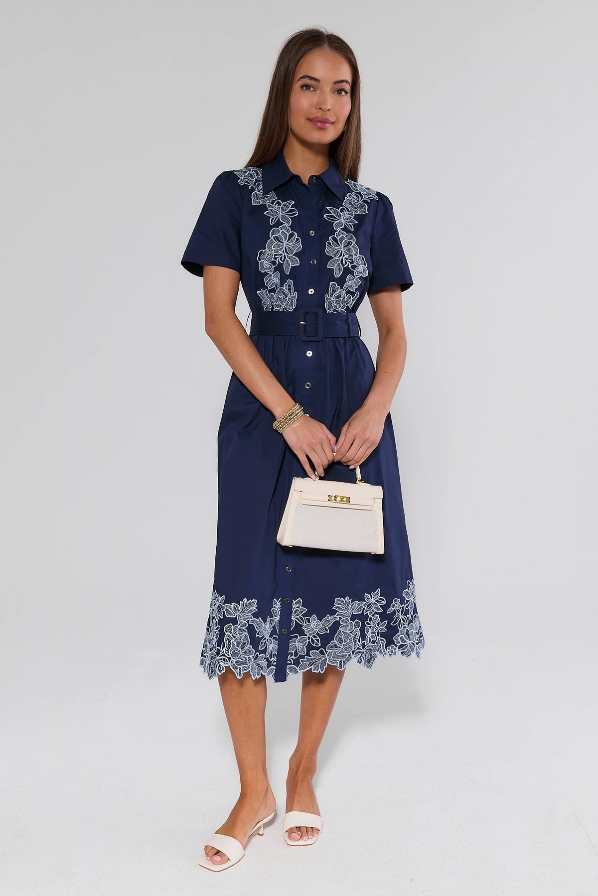 Woman wearing navy embroidered midi dress with white floral lace detail, belted waist, holding cream handbag - Front View | Avara