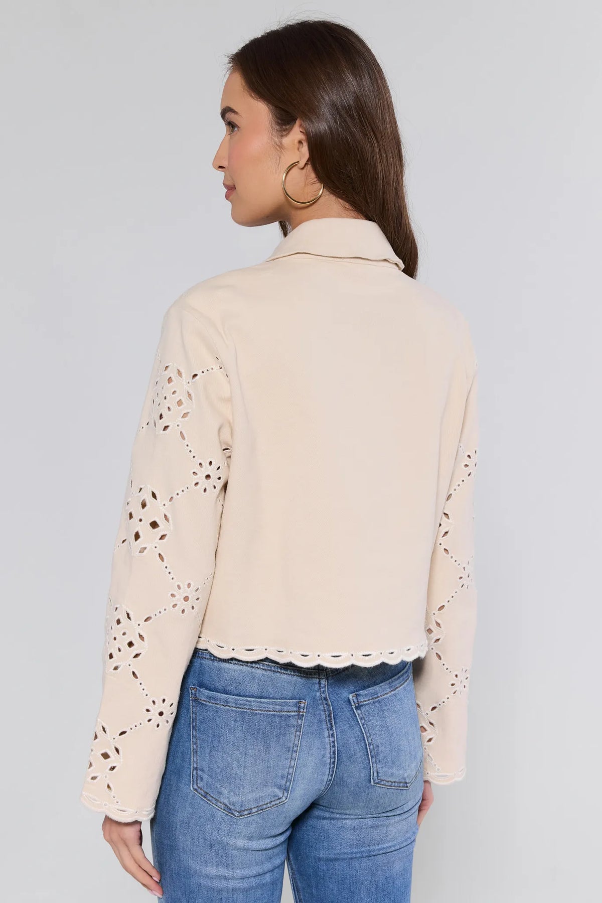 Person wearing Gina Beige Eyelet Jacket featuring geometric-floral lace sleeves, scalloped hem, and relaxed fit, paired with medium-wash jeans, seen from behind.