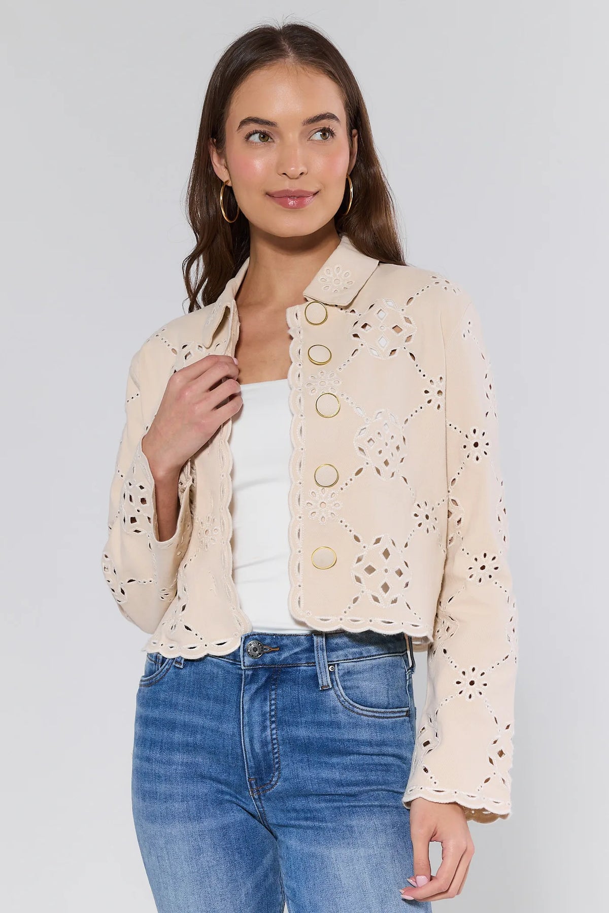 Woman models the Gina Beige Eyelet Jacket, featuring scalloped edges, geometric cut-outs, gold-tone grommet buttons, and a cropped, relaxed fit over a white tank and jeans.