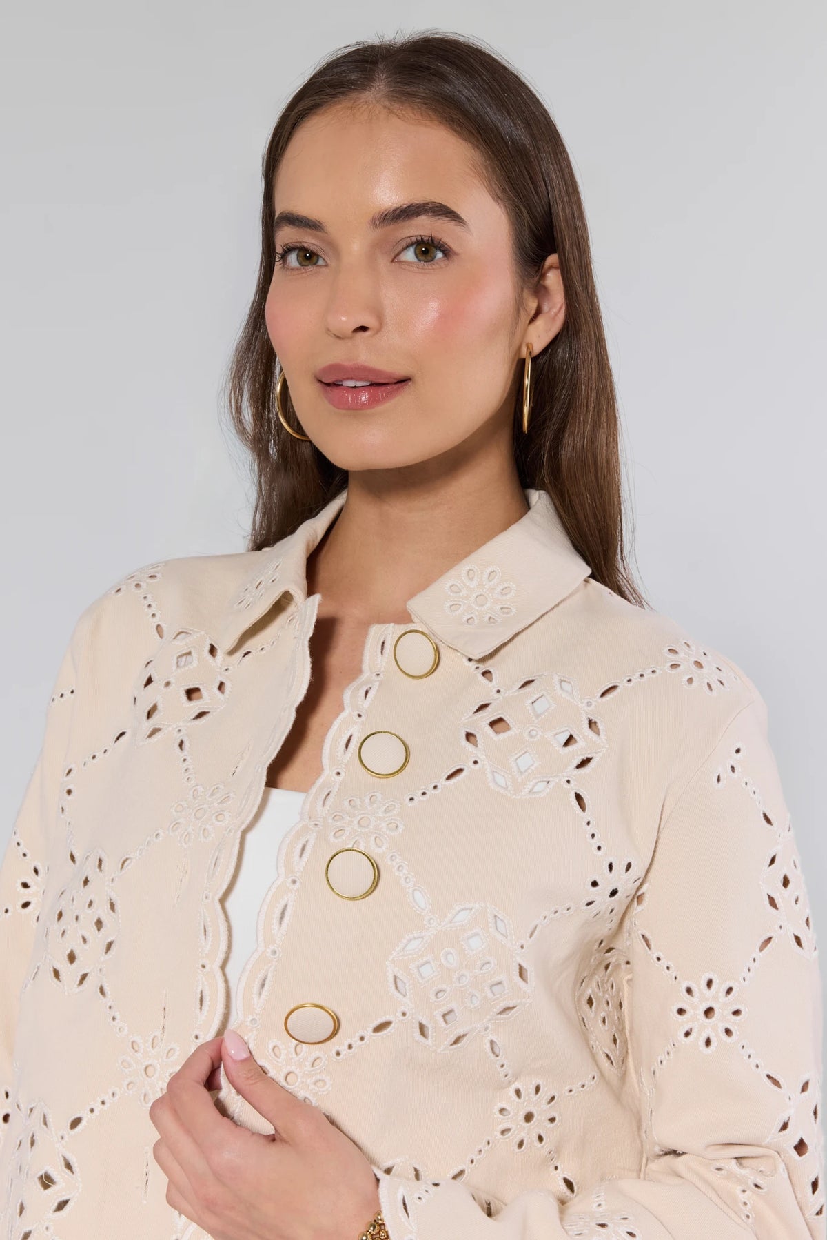 Woman modeling the Gina Beige Eyelet Jacket with broderie anglaise, scalloped edges, and gold-tone ring closures, showcasing a relaxed, cropped fit and lightweight design.