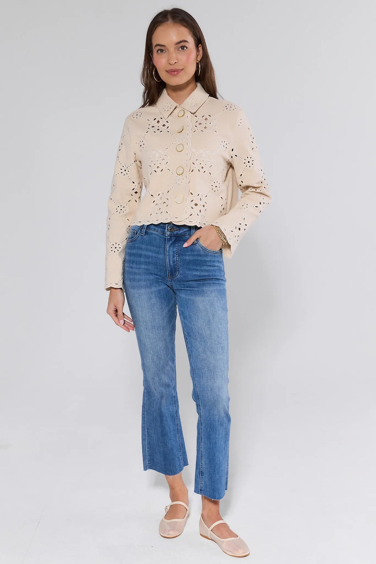 Young woman models Gina Beige Eyelet Jacket with gold buttons, scalloped trim, and a cropped hem over jeans. Paired with Mary Jane flats and gold jewelry.