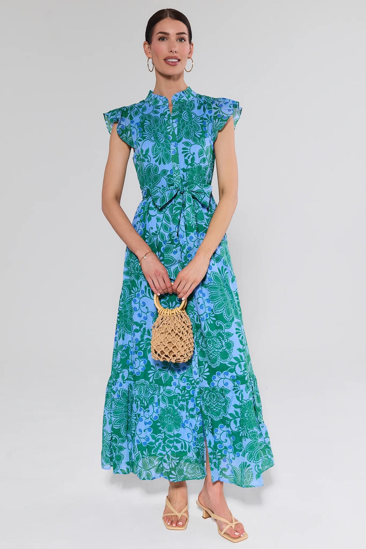 Woman wearing a green and blue floral dress holding a woven bag on a white background