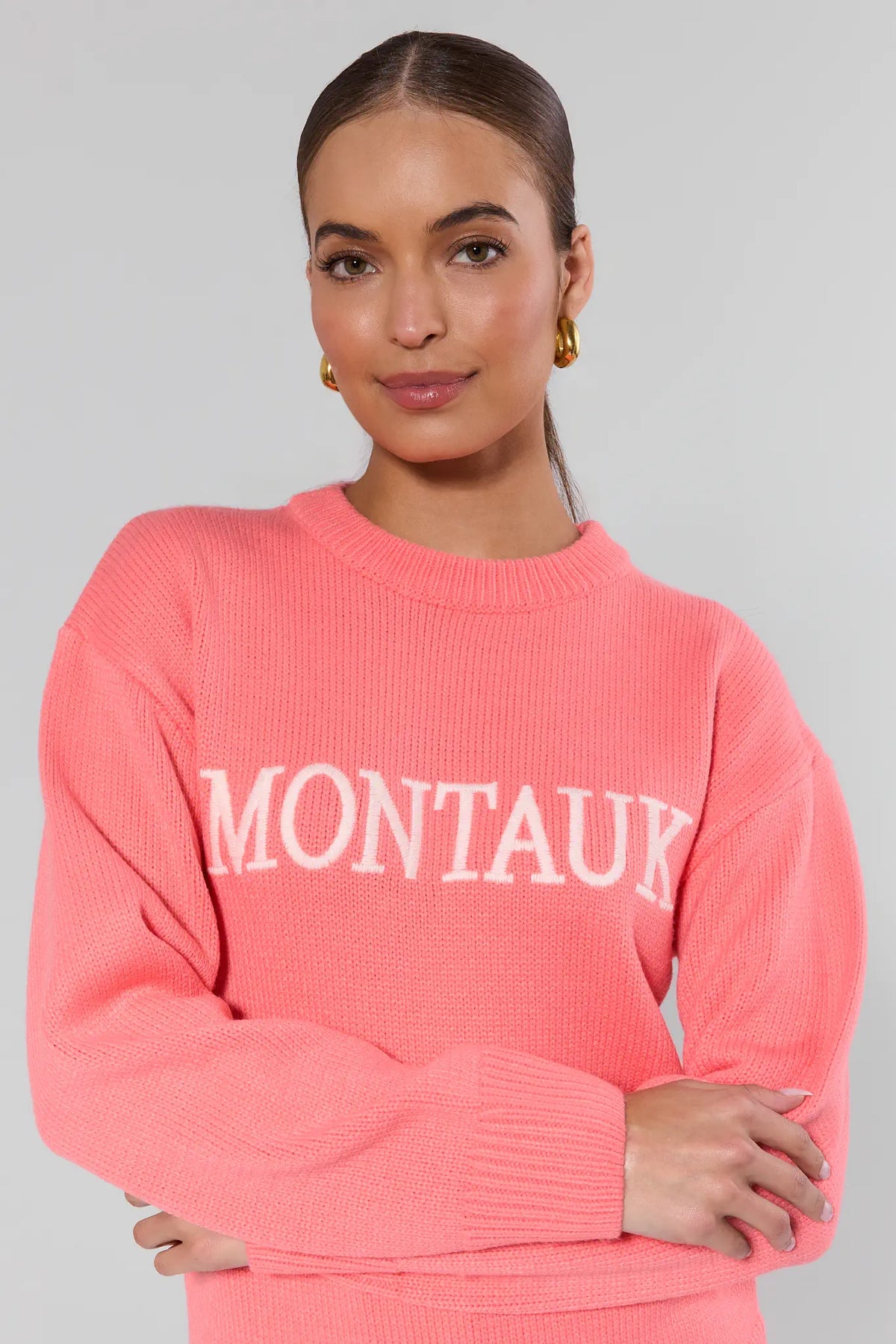 Montauk coral knit sweater close-up showing contrast letter detail and ribbed cuffs, arms crossed | Avara