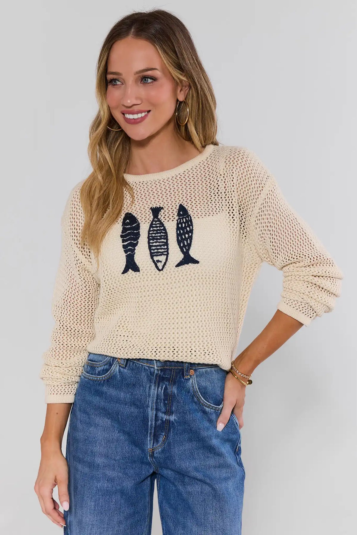 Woman wearing a beige knit sweater with fish designs and blue jeans on a plain background