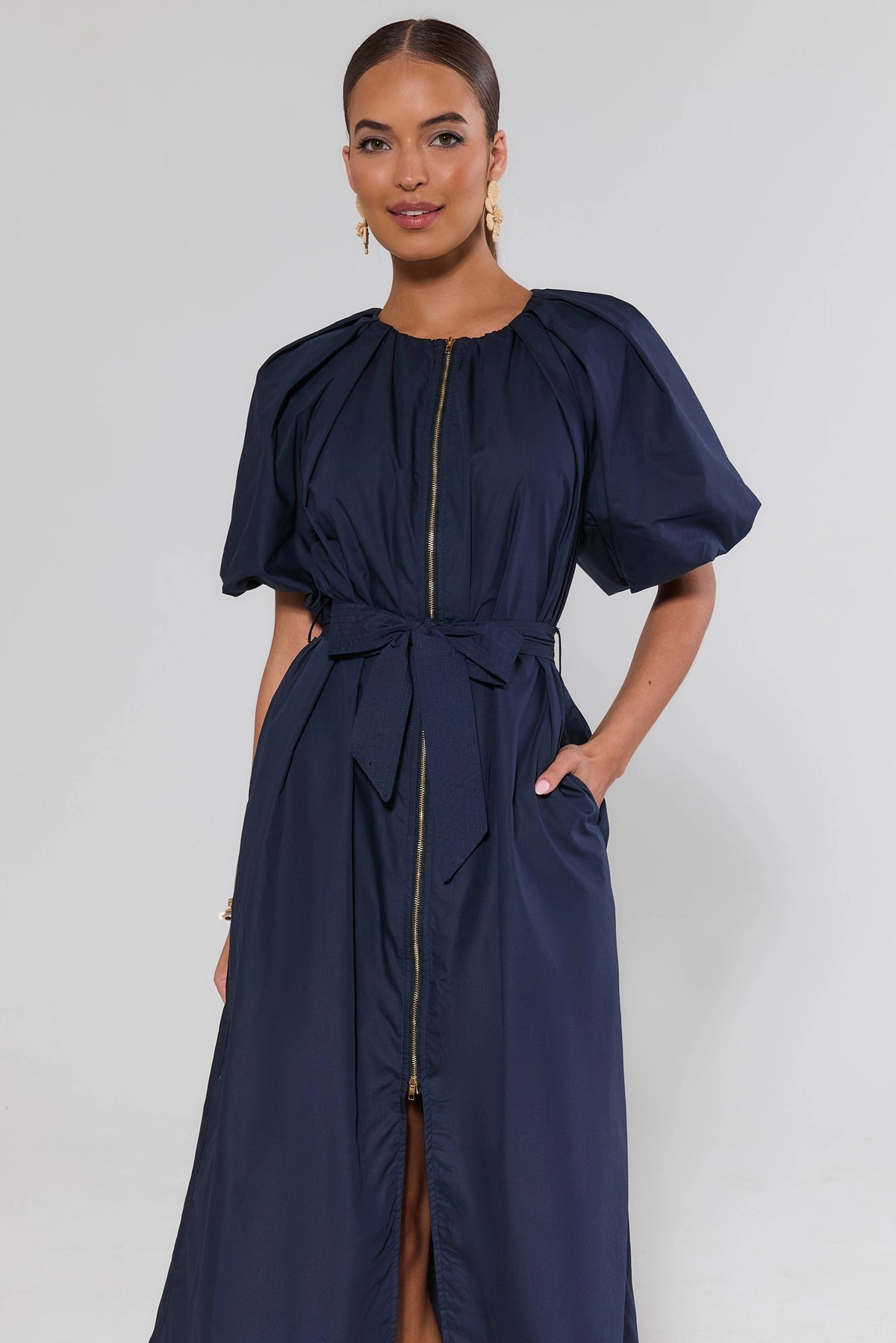 Woman wearing navy belted midi dress with puff sleeves and gold zip front, three-quarter view | Avara