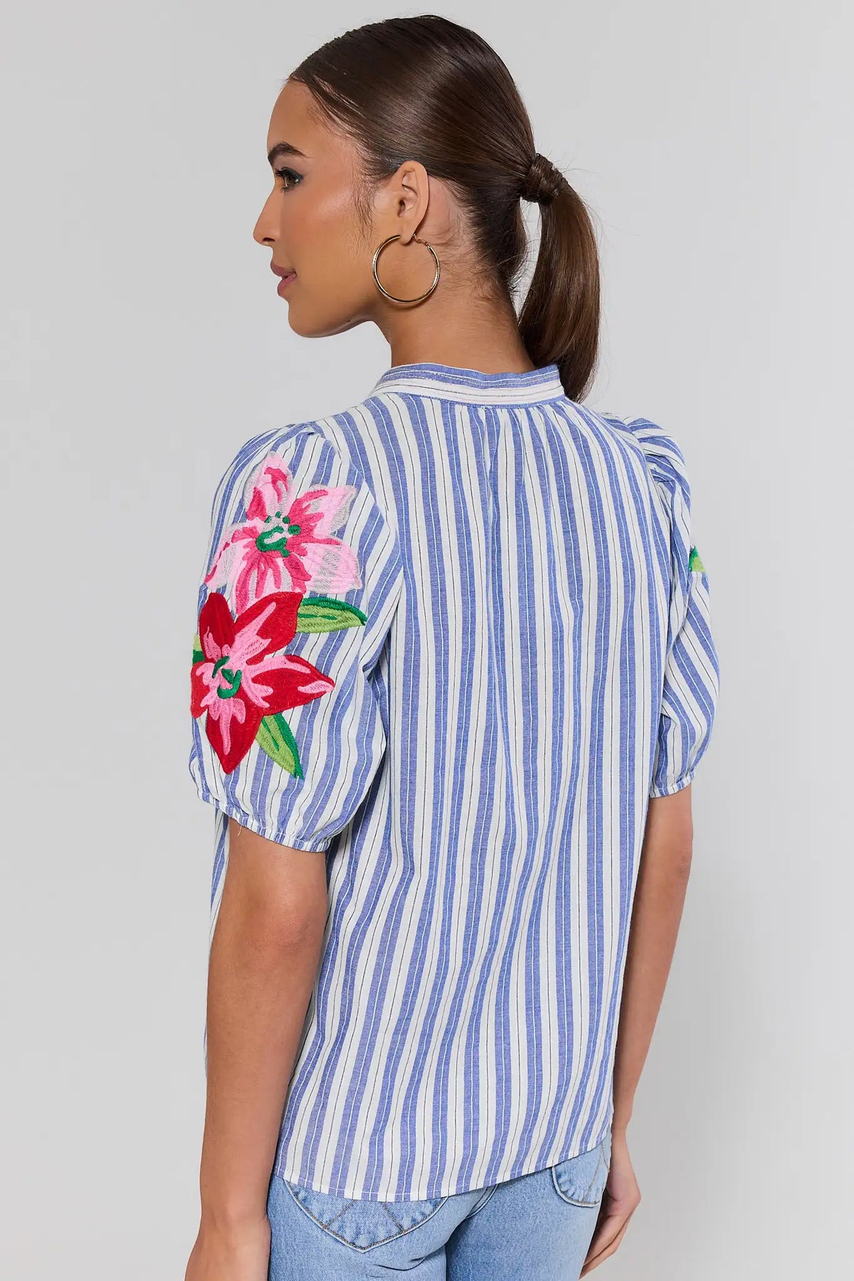 Woman standing wearing a blue and white striped shirt with floral embroidery on a plain background