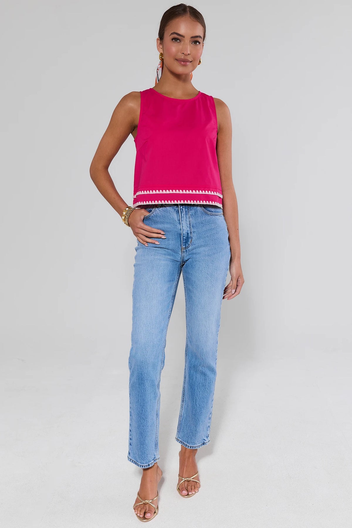 Woman wearing a pink sleeveless top with white trim and blue jeans on a gray background