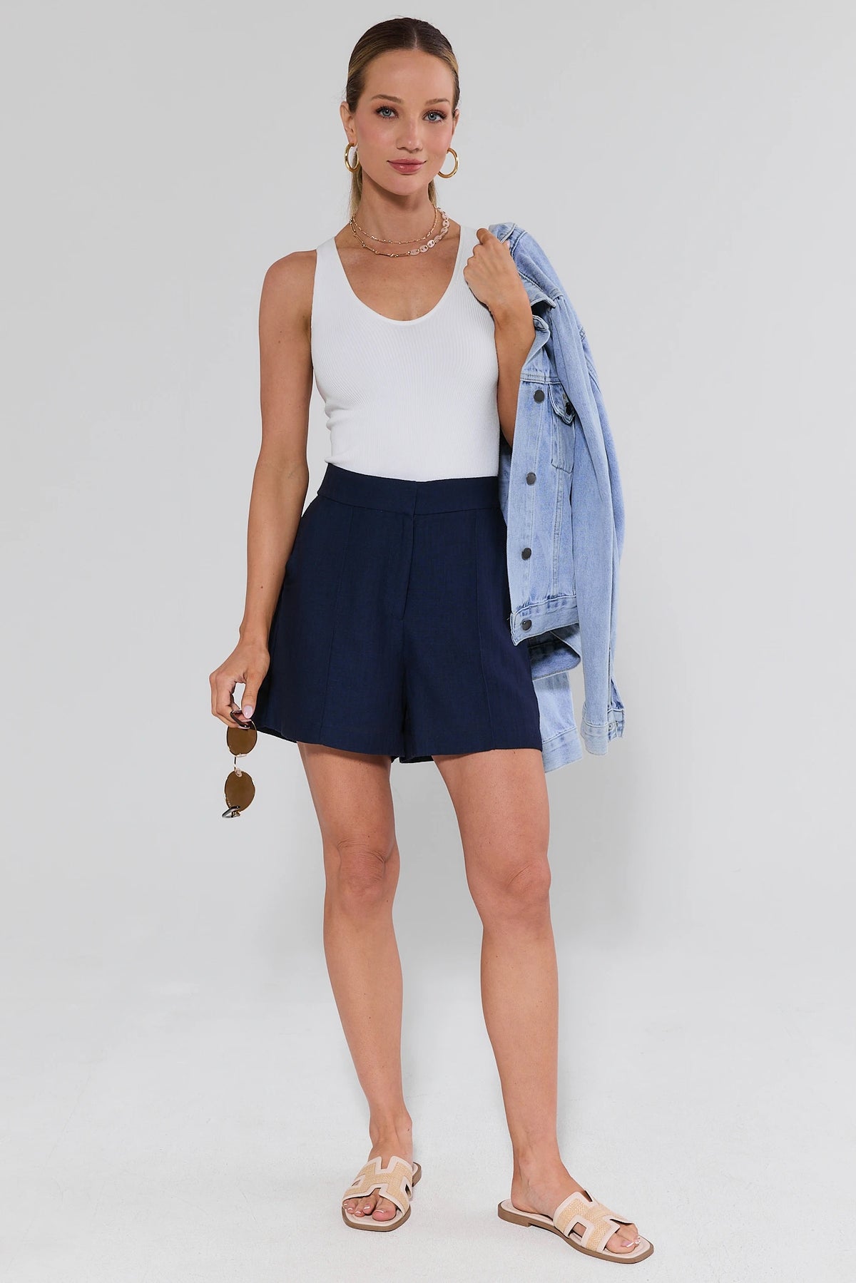 Woman wearing a white tank top and navy shorts with a denim jacket over her shoulder on a plain background
