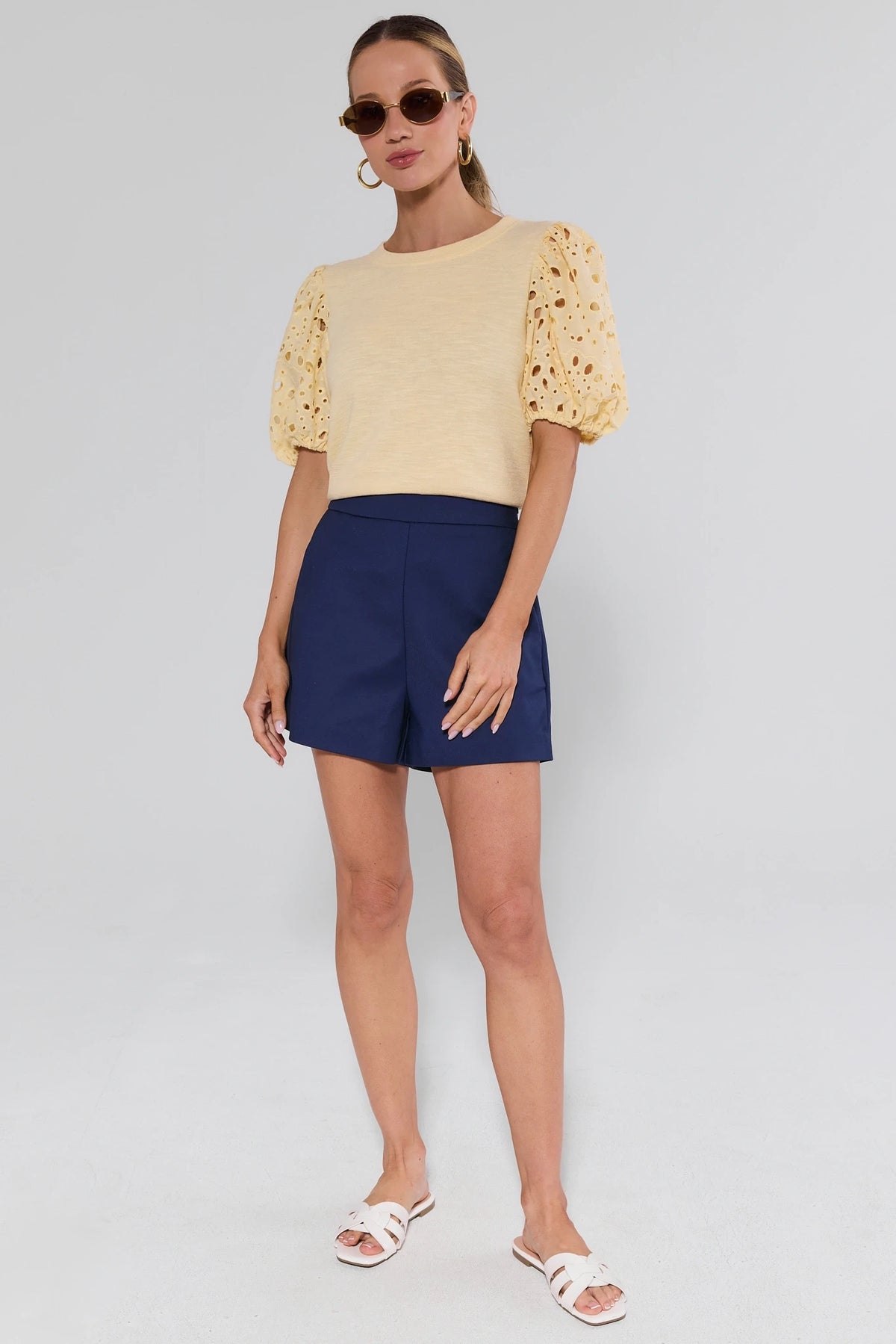 Woman wearing a yellow top with lace sleeves and navy shorts on a gray background