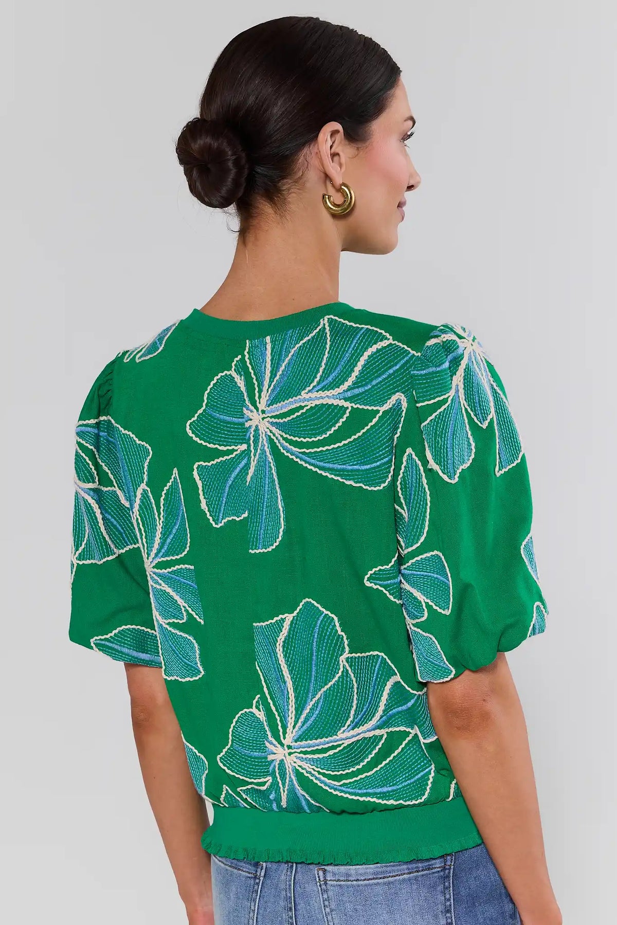 Woman standing wearing a green blouse with floral patterns on a plain background