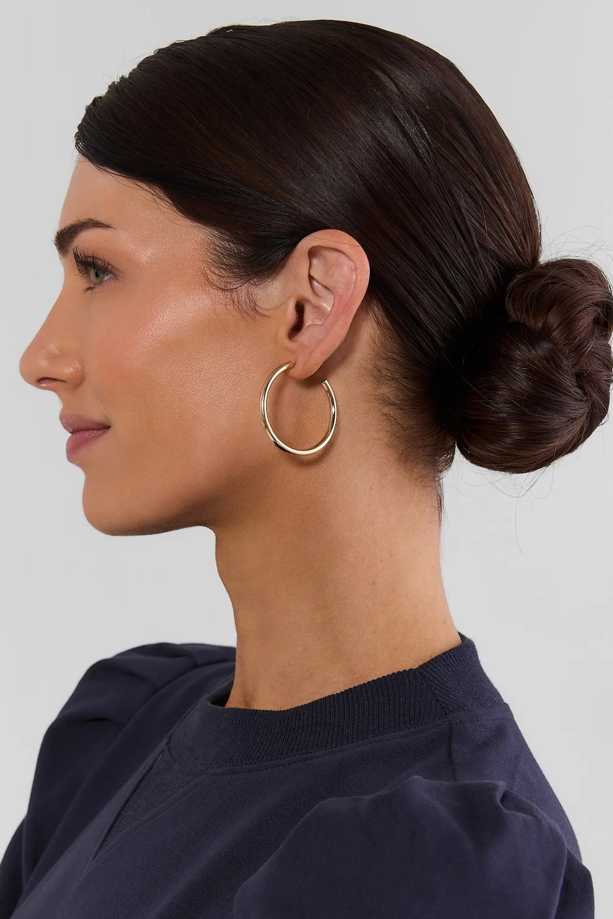 Woman in profile wearing Emily Gold Medium Hoop Earrings, low bun, navy top - On Model | Avara