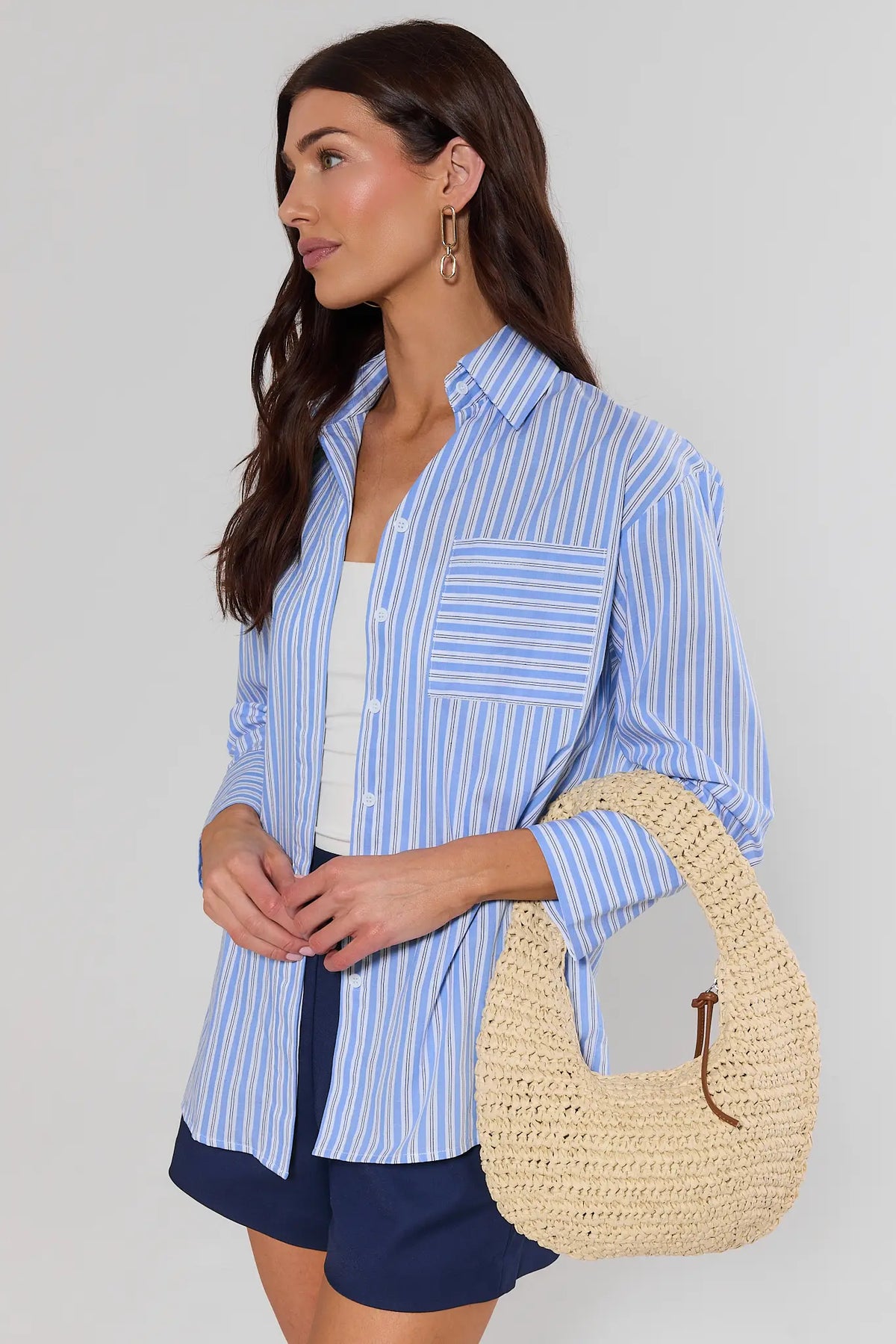 Woman wearing a blue striped shirt and navy shorts, holding a straw bag against a plain background