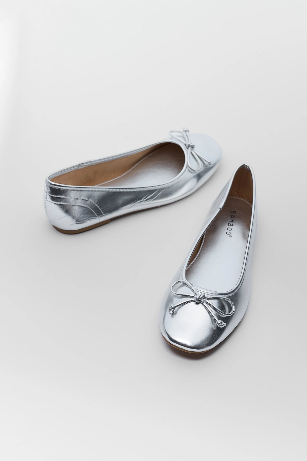 Silver bow ballet flats pair shown at angle, metallic finish with round toe and tan insole visible | Avara