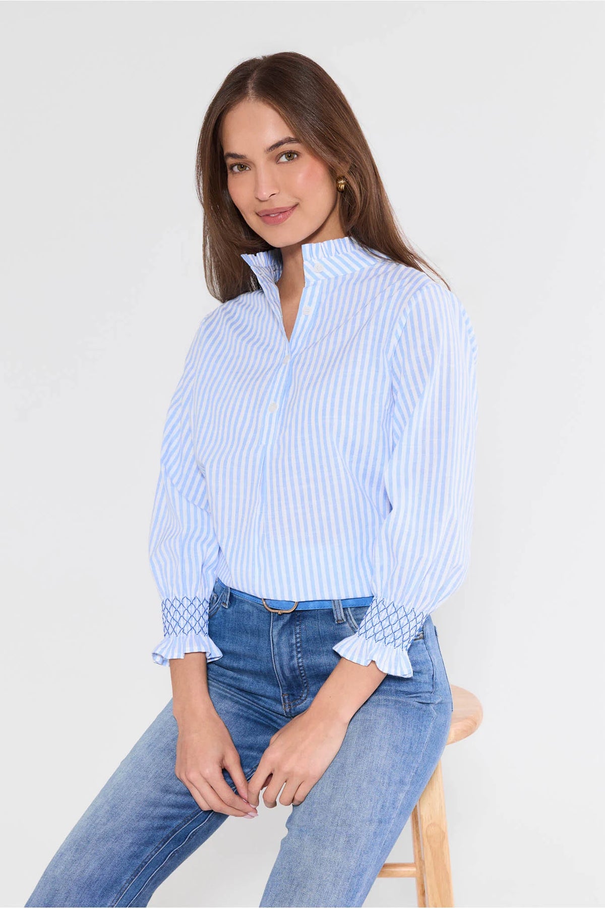 Woman wearing a light blue striped shirt and jeans sitting on a stool against a white background