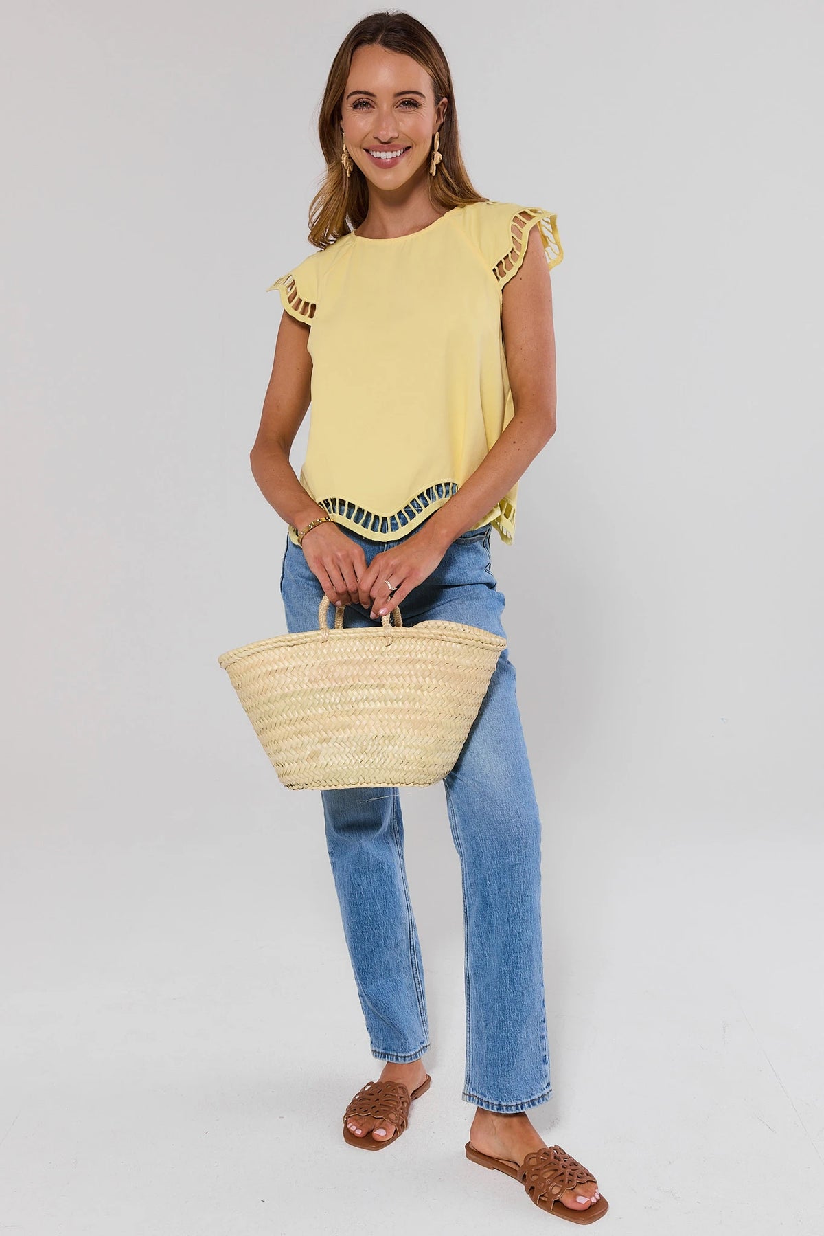 Woman standing wearing a yellow top and blue jeans, holding a straw bag on a plain background