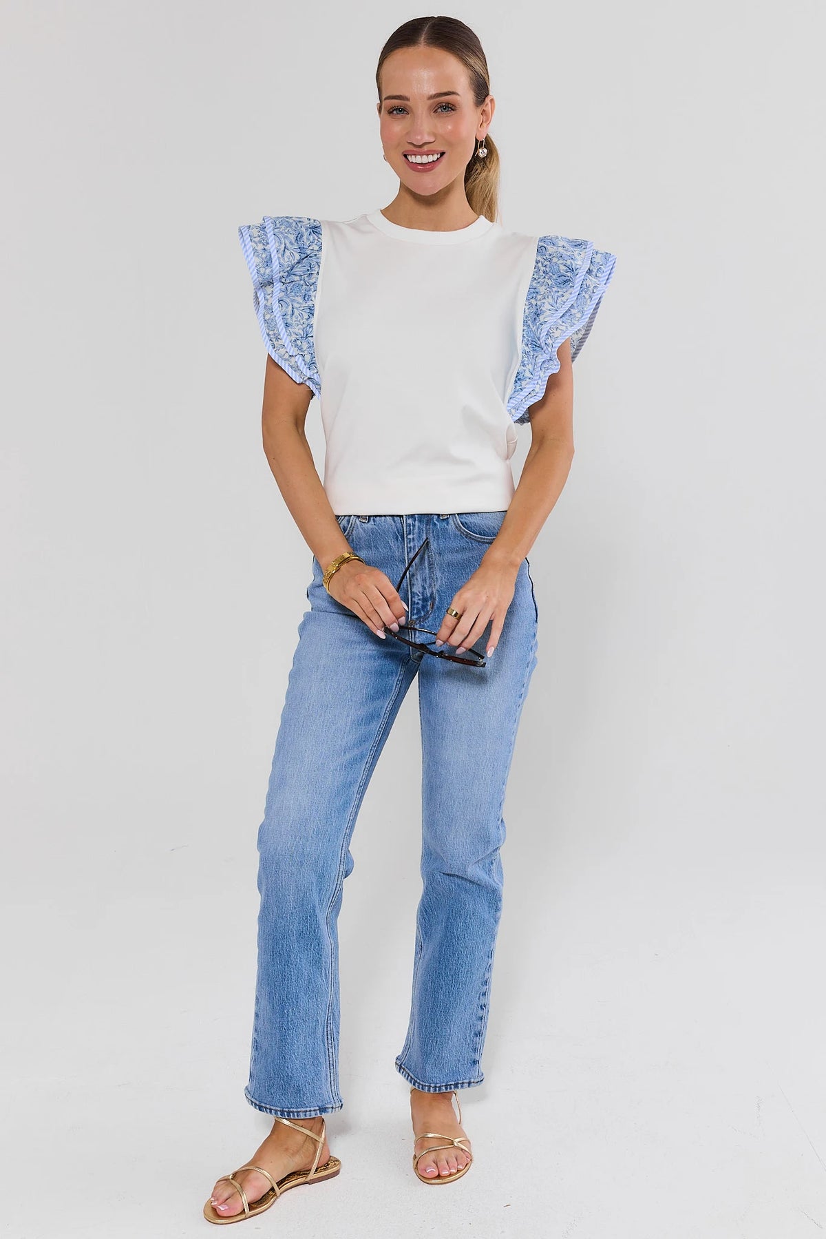 Woman wearing a white top with blue ruffled sleeves and light blue jeans on a white background