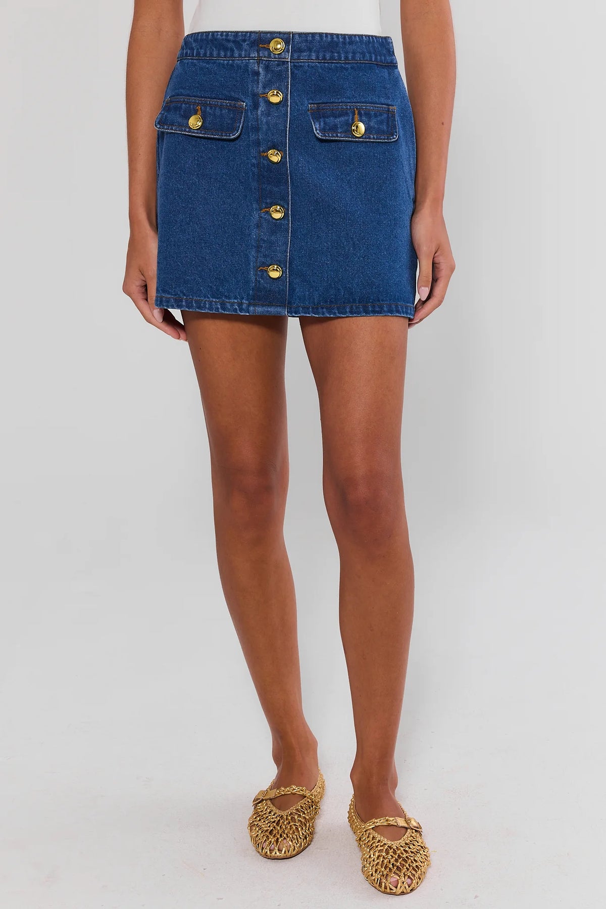 Model wearing blue Vickie Denim Button Up Skirt with gold buttons, paired with woven gold flats - Front View | Avara