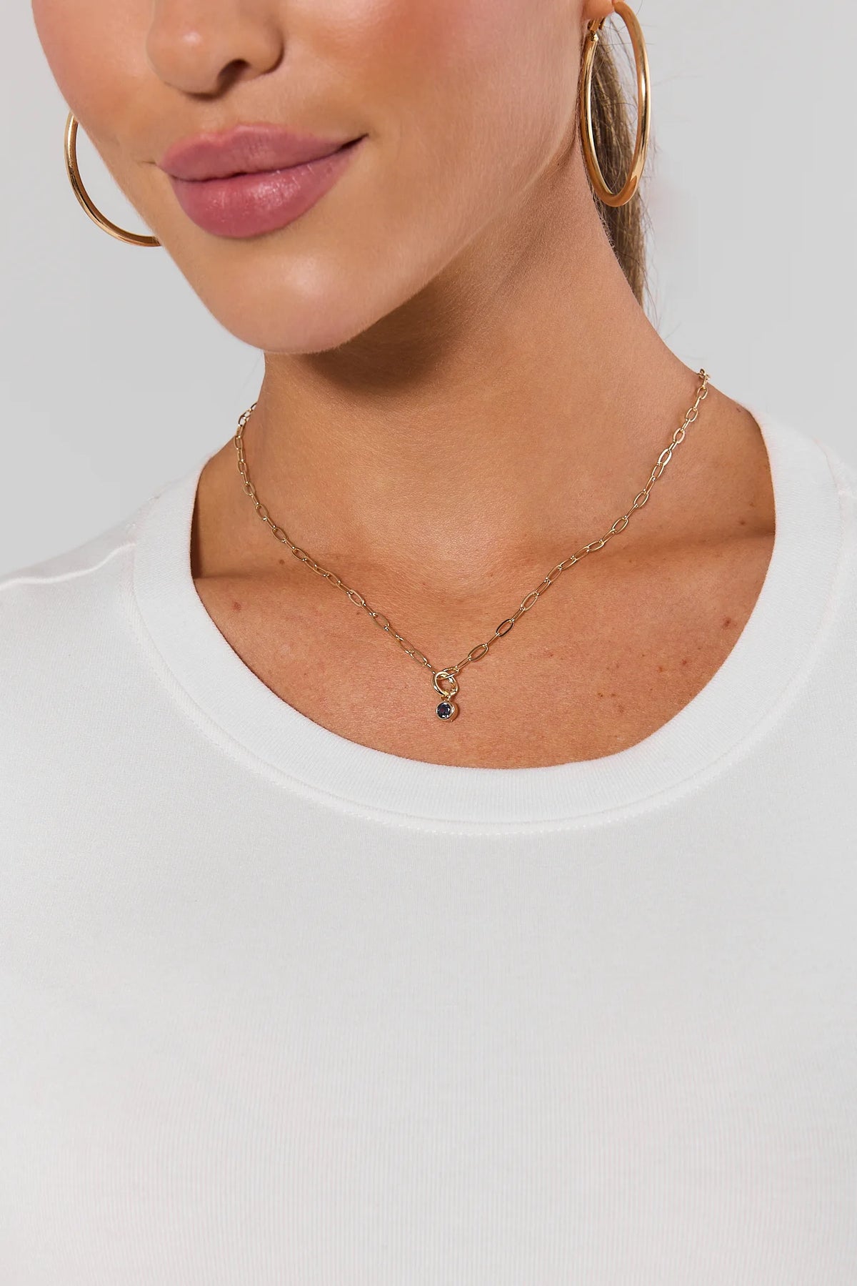 Purple rhinestone charm on gold chain necklace styled on model with white top and gold hoops | Avara
