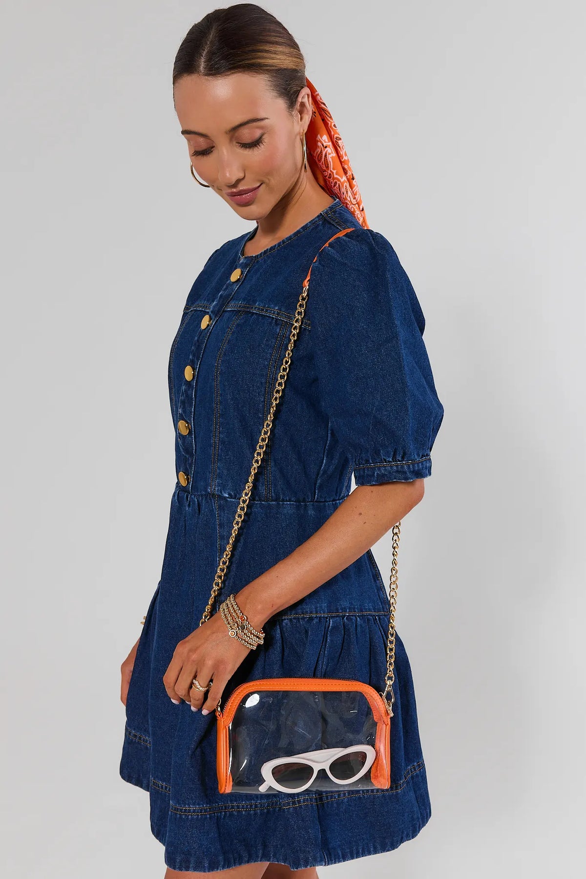 Woman in blue denim dress carrying orange trim clear crossbody bag with gold chain — game day styled look | Avara