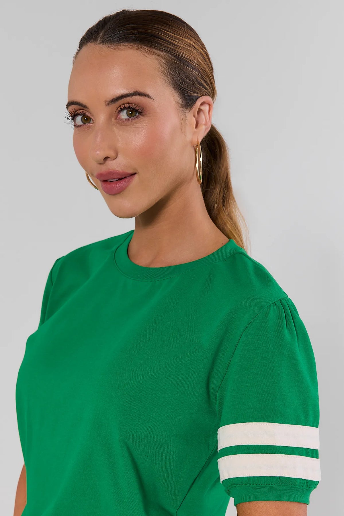 Close up of a woman wearing a bright green short-sleeve crewneck top with two white stripes on the sleeve, shown against a plain light gray studio background.