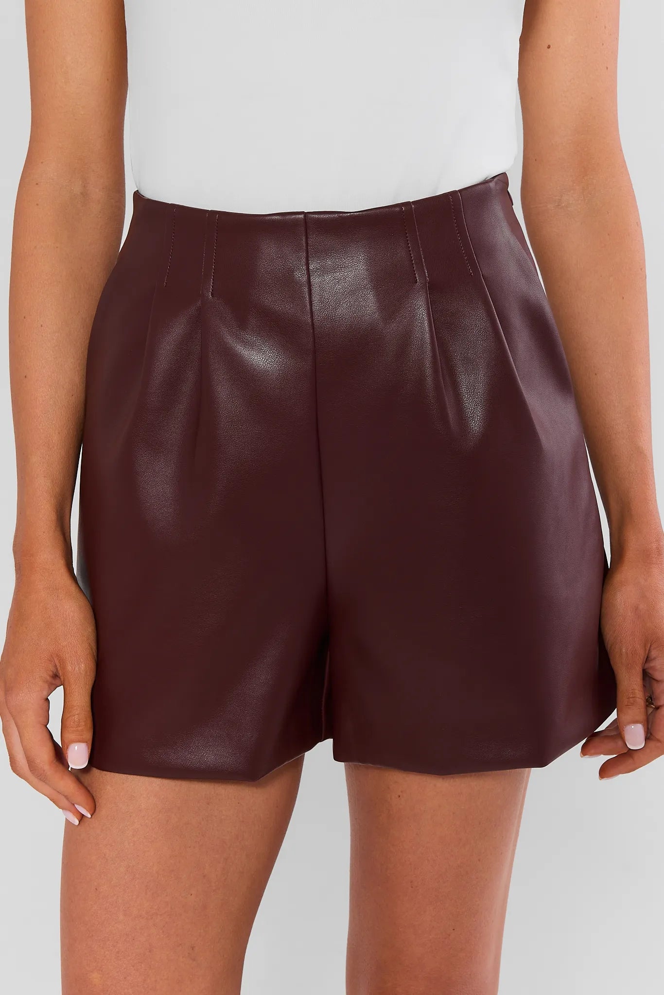 High-waisted burgundy faux leather shorts with front pleats, close-up front view on model | Avara