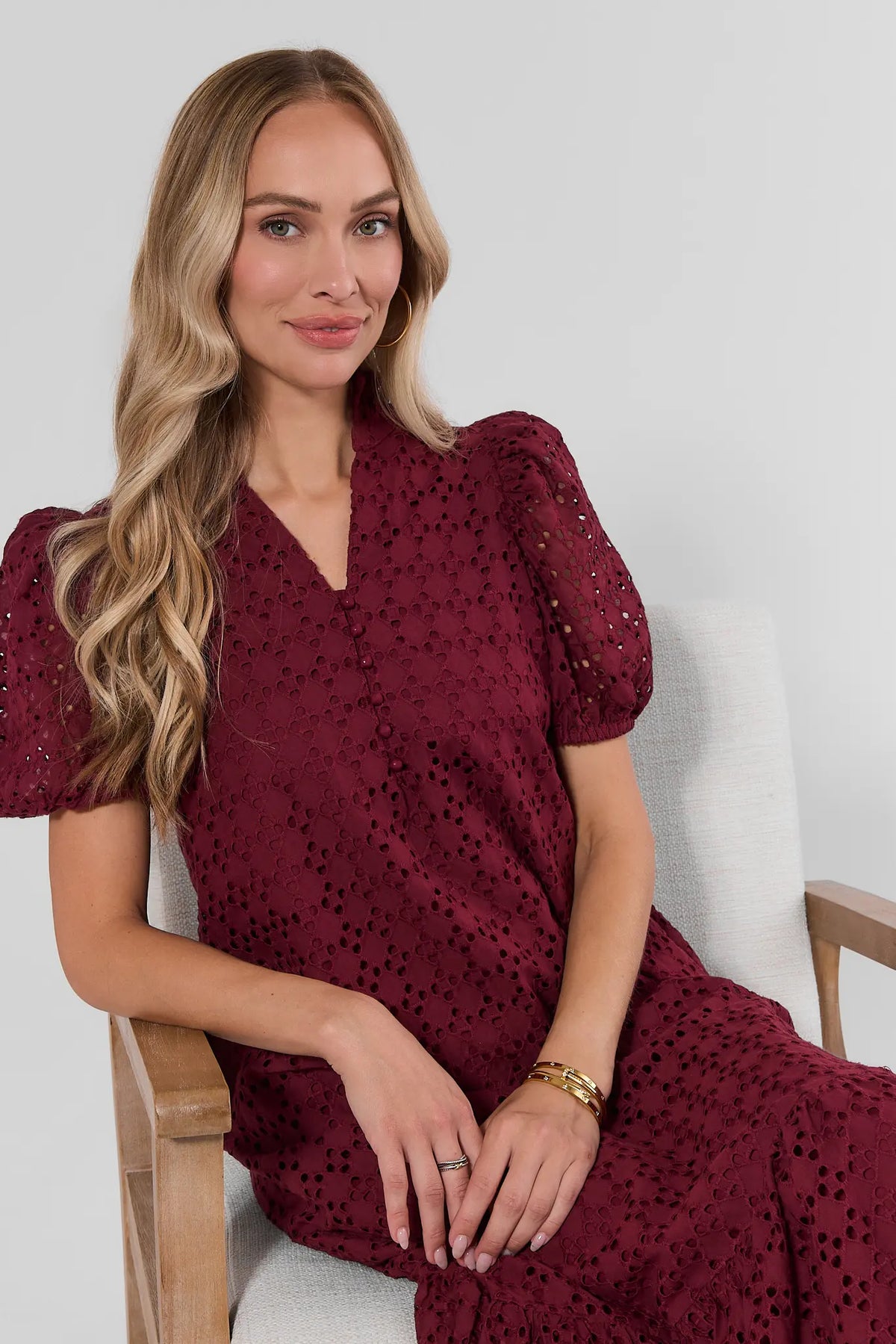 Burgundy eyelet maxi dress seated lifestyle shot, puff sleeves and V-neckline, gold bracelet styling | Avara