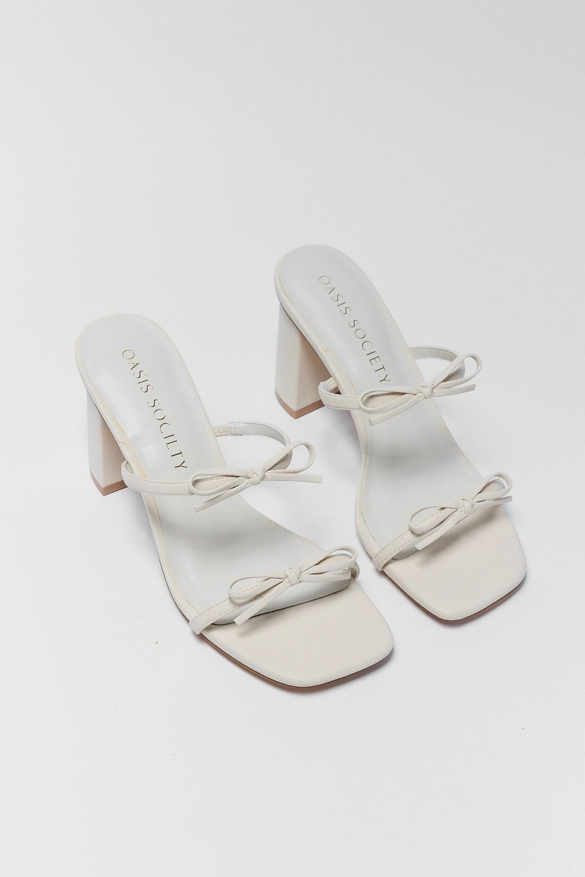 White double-strap bow block heels pair, angled flat lay showing toe and heel | Avara